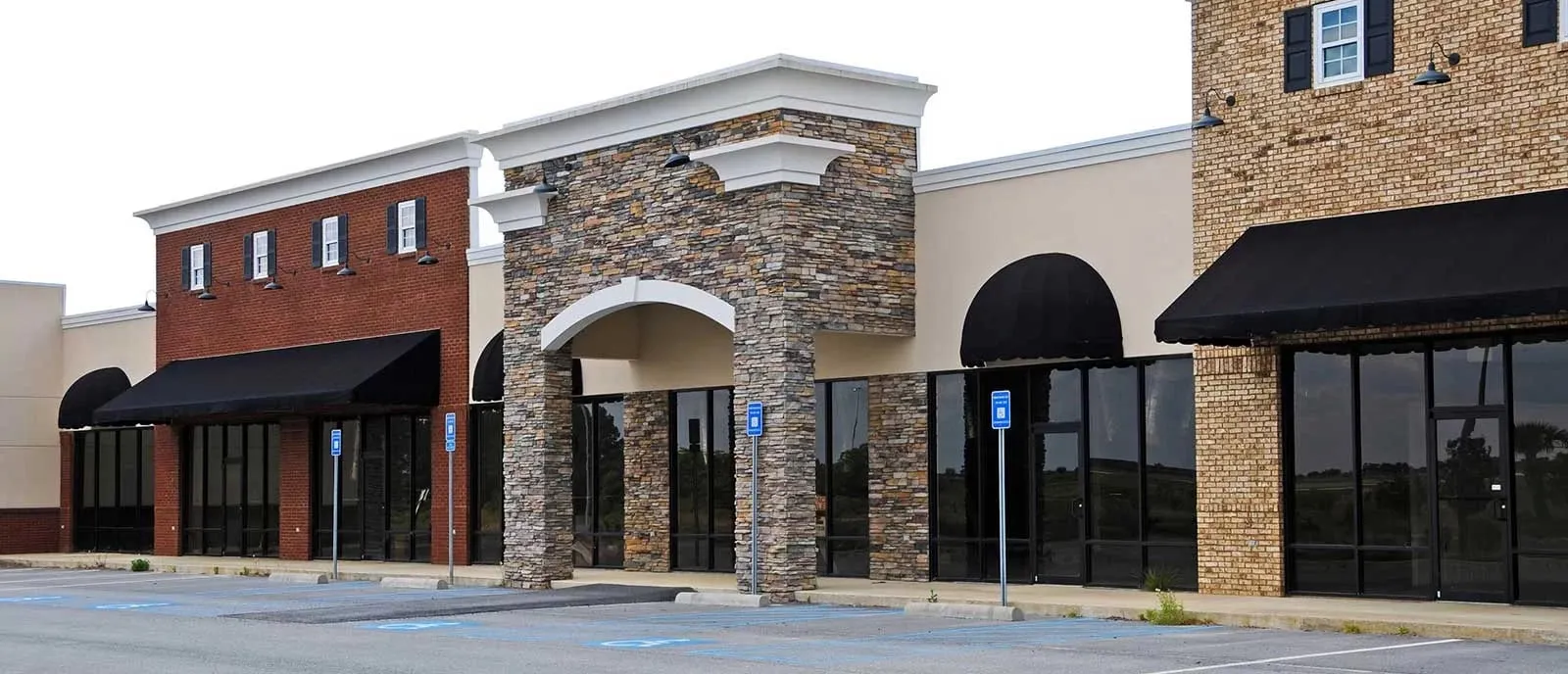 A commercial retail plaza with storefronts, awnings, and an empty parking lot.