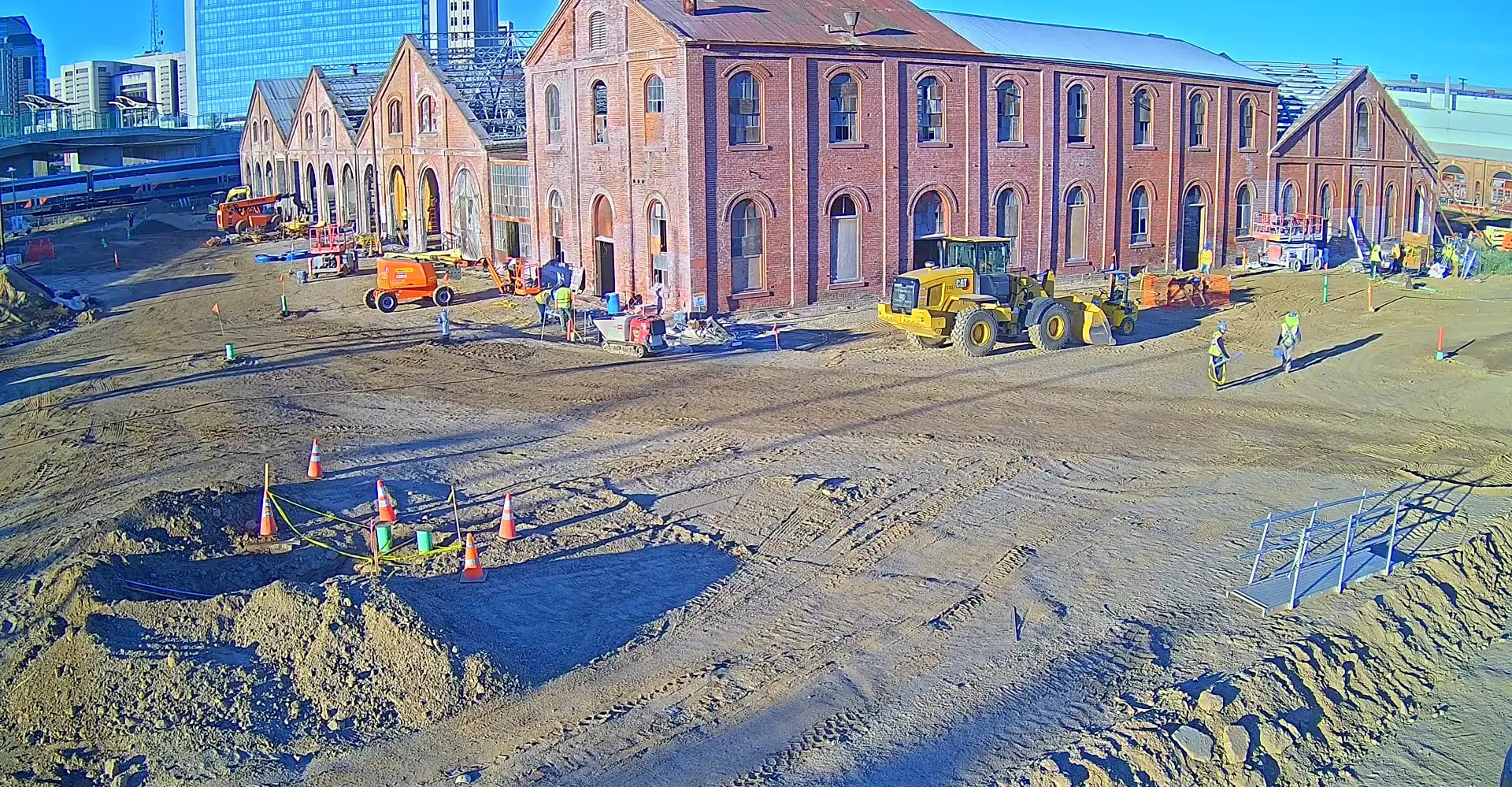 An Odin Systems site-security camera view of an active construction site — graded dirt, heavy equipment, and crews working beside historic brick buildings.