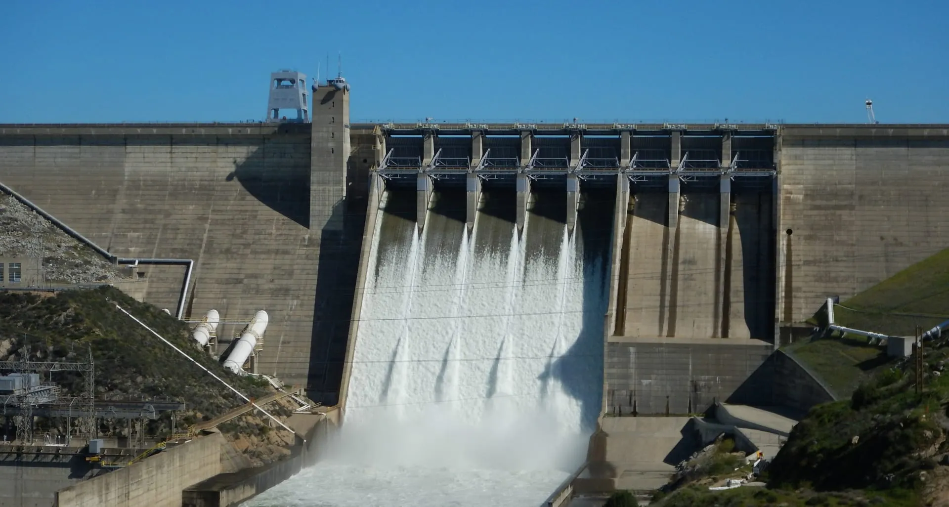 Water releasing through the spillway gates of a large concrete dam under a clear blue sky.