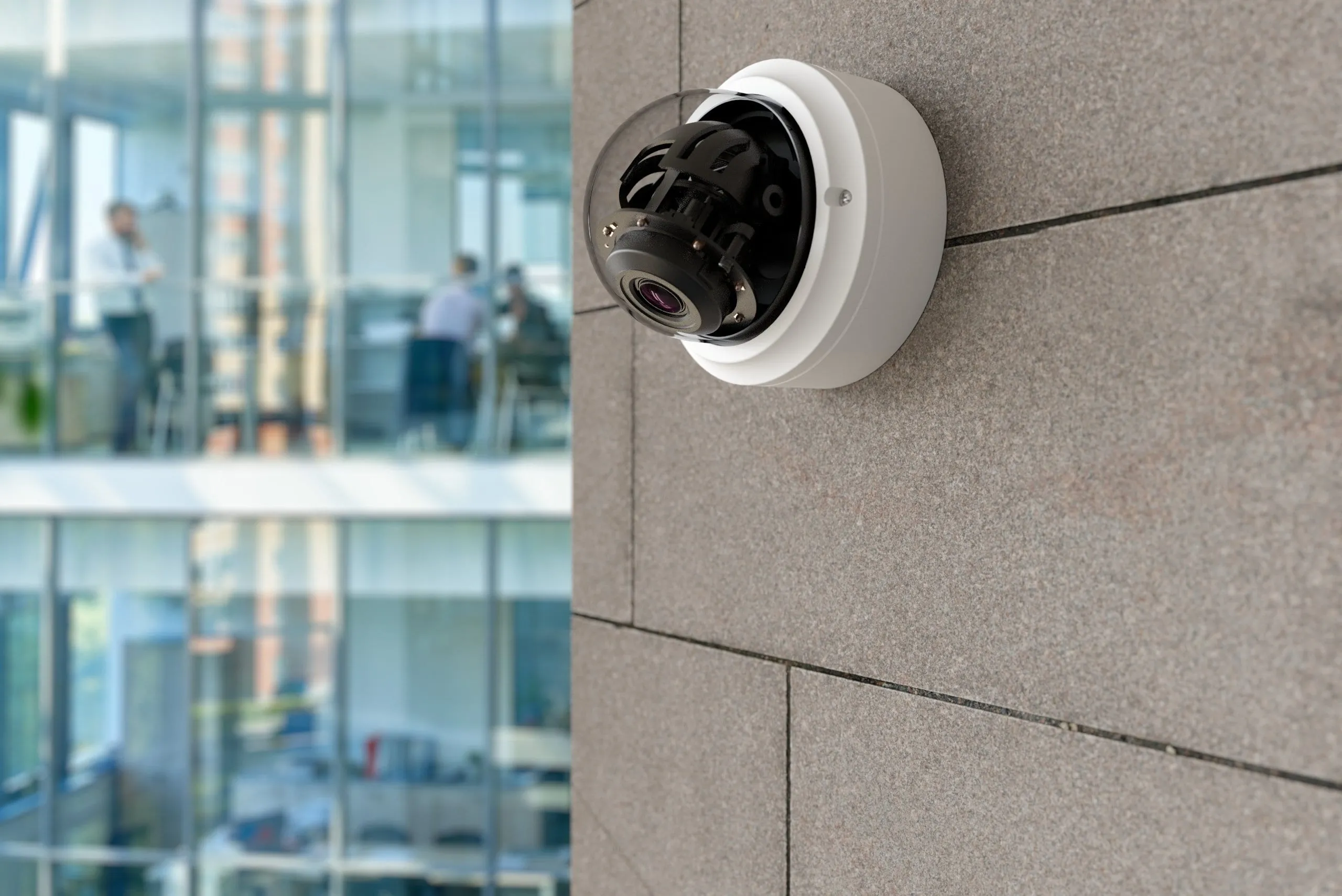 A fixed dome security camera mounted on the exterior wall of a modern glass office building — Odin Systems designs and installs permanent IP video security systems across California.