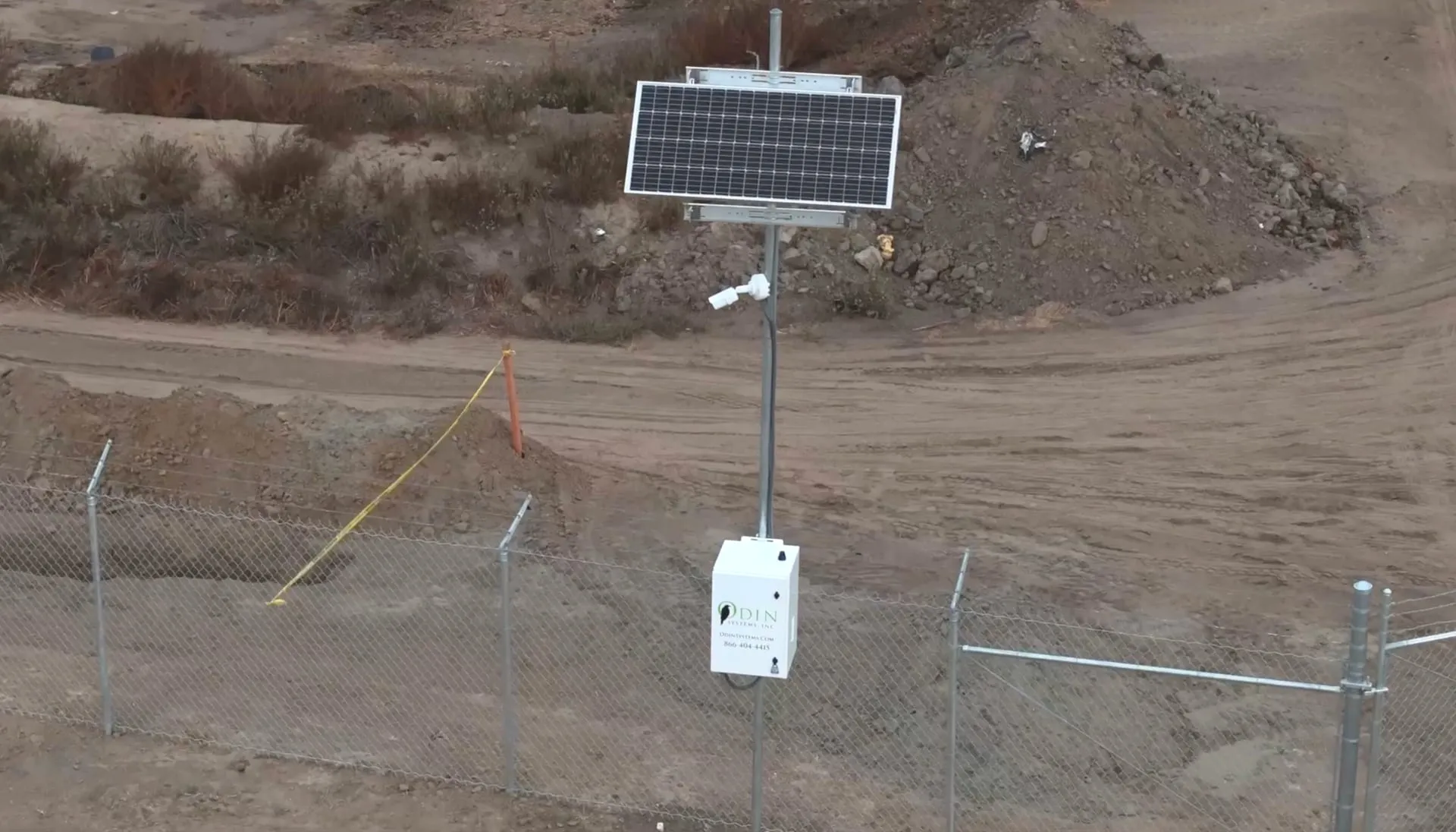 An Odin Systems Solar RVN Pod — a pole-mounted solar panel, camera, and Odin-branded enclosure — deployed behind a chain-link fence at a graded construction site.