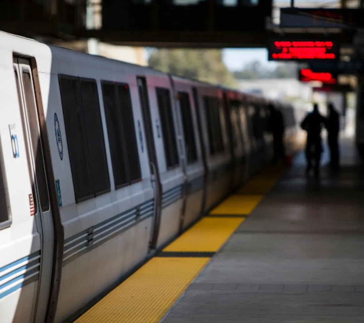 A BART commuter train at a station platform, with riders waiting along the yellow platform edge.
